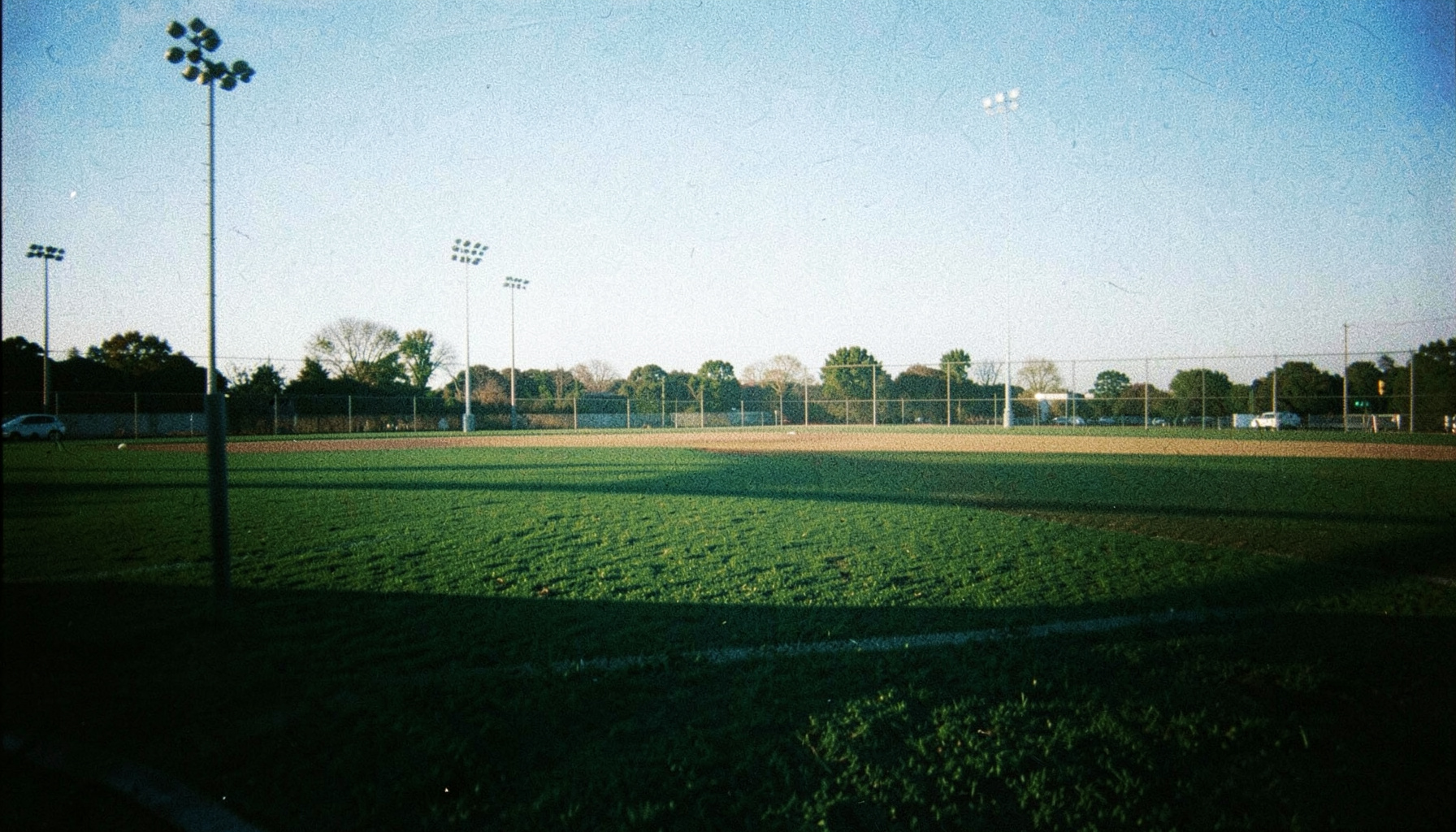 A grainy film photograph of a baseball diamond seen from behind the center field fence, late-afternoon light casting long shadows across the outfield grass, deep green turf against a pale blue sky.
